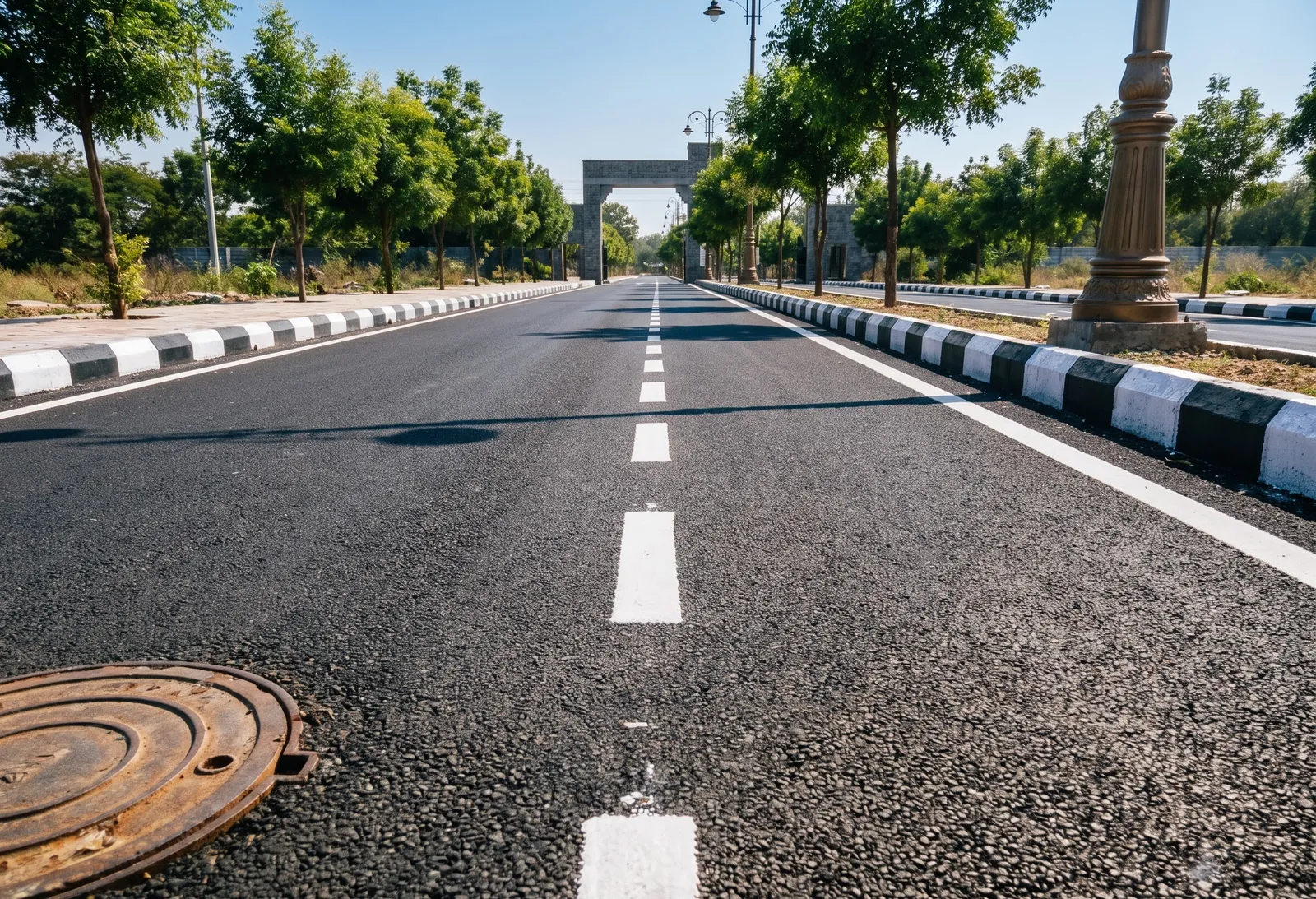 A new black-top road through a plotted layout with kerbs, street lights and young trees